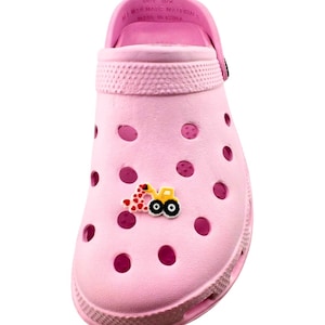 May include: A light pink clog shoe with a decorative yellow and red tractor charm. The shoe has multiple ventilation holes and a pivoting heel strap. The text "SIZE 5/6" and "MAN MADE MATERIALS MADE IN CHINA" is visible on the strap.