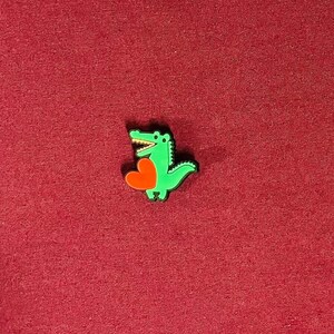 May include: A green alligator charm holding a red heart. The alligator has a white belly and a black outline. The charm is on a red textured background.