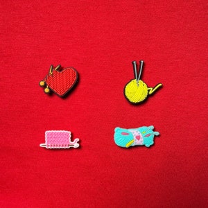 May include: Four colorful knitting-themed charms. A red heart with knitting needles, a yellow ball of yarn with needles, a pink knitted rectangle with a hook, and a turquoise yarn roll with a pink heart.