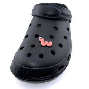 May include: Black clog-style shoe with a pink worm-shaped charm. The shoe has multiple ventilation holes and a textured sole. The charm is a light pink color with a darker pink outline. The shoe is made of a rubbery material.