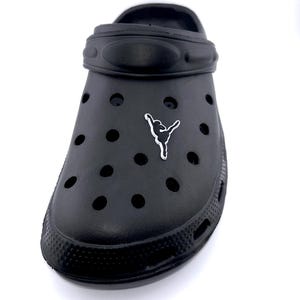 May include: Black clog-style shoe with multiple ventilation holes and a pivoting heel strap. A small, white graphic of a person in a ballet pose is affixed to the side of the shoe. The shoe is made of a rubber-like material.