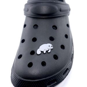 May include: Black clog shoe with a white manatee charm. The shoe has multiple ventilation holes and a textured sole. The manatee charm is a simple outline with a smiling face. The shoe is made of a rubbery material.