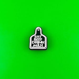May include: Black and white enamel pin shaped like a cow tag with the text "EAT BEEF DRINK MILK".