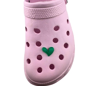 May include: A light pink clog-style shoe with multiple round ventilation holes. A green heart-shaped charm is attached to the front. The shoe has a textured band around the top and is made of a rubbery material.
