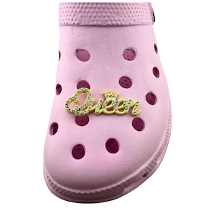 May include: A light pink clog shoe with multiple round holes. The word "Queen" is written in gold-colored script with rhinestone accents. The shoe has a textured surface and a rounded toe.