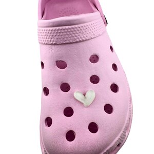 May include: A light pink clog shoe with multiple ventilation holes and a white heart-shaped charm. The shoe has a textured surface and a rounded toe. The text "SIZE 5/6" is visible on the strap.