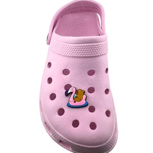 May include: A light pink clog shoe with a decorative charm featuring a flamingo and a bear on a float. The shoe has multiple ventilation holes and a pivoting heel strap. The text "SIZE 5/6 ALL MAN MADE MATERIAL MADE IN CHINA" is visible on the strap.