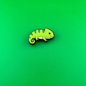 May include: A green and black cartoon chameleon charm with a smiling face.
