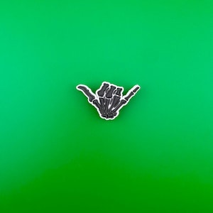 May include: A black skeleton hand making the shaka sign, a popular Hawaiian gesture, against a bright green background.