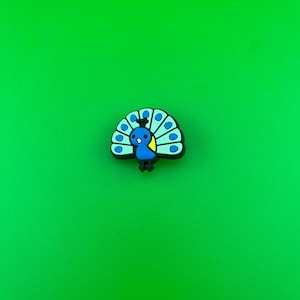 May include: A blue and green peacock charm with a yellow beak and white eyes. The charm is made of rubber and has a flat back.