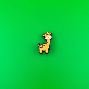 May include: A small, cartoon giraffe figurine with brown spots on a yellow body. The giraffe has a black outline and a white face with pink cheeks. The figurine is on a green background.