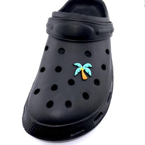 May include: Black clog shoe with a palm tree charm. The shoe has multiple round holes and a textured sole. The palm tree charm is in shades of blue, green, and orange. The shoe is made of a rubber-like material.
