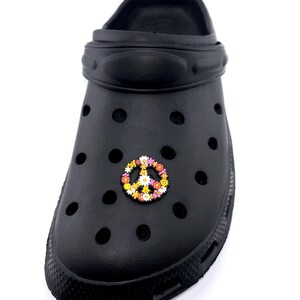 May include: Black clog shoe with a colorful peace sign charm. The charm features a peace symbol made of small flowers in shades of yellow, pink, orange, and white. The shoe has multiple ventilation holes.