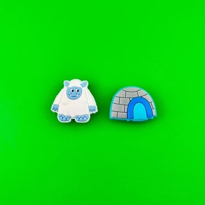 May include: A white yeti with blue eyes and a blue mouth, and a gray igloo with a blue door on a green background.