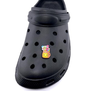 May include: Black clog-style shoe with a decorative charm. The charm features a yellow bag with colorful art supplies. The shoe has multiple ventilation holes and a pivoting heel strap. The shoe is made of a rubber-like material.