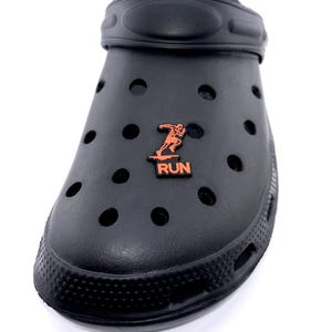May include: Black clog-style shoe with multiple ventilation holes. An orange emblem of a running figure and the word "RUN" is affixed to the shoe. The shoe is made of a rubbery material and is designed for casual wear.