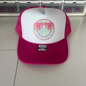 May include: A pink and white trucker hat with a white smiley face with pink sunglasses and a palm tree design. The hat has a black label that says "OTTO WORLD FAMOUS FOAM FRONT TRUCKER".