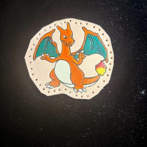 May include: Hand-painted leather patch featuring a Charizard design. The orange dragon has green wings and a flame at the tail. The patch is beige with a stitched border and is suitable for jackets, bags, or other accessories.