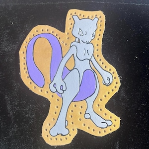 May include: A hand-painted, light gray and purple Mewtwo character design on a tan leather patch. The patch has a decorative border with small, evenly spaced holes. The background is black.