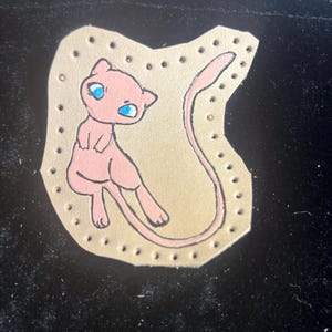 May include: A pink cartoon character with blue eyes is painted on a tan leather patch. The character has a long tail and is surrounded by a border of small holes. The patch is on a black surface.