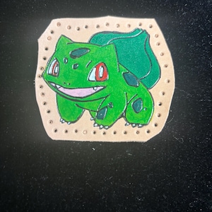 May include: A green Bulbasaur character illustration on a tan leather patch. The Bulbasaur has red eyes, black spots, and a smiling mouth. The patch is surrounded by small, evenly spaced holes.