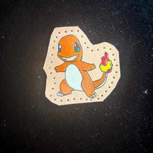 May include: Hand-drawn orange Charmander character on a tan leather patch. The cartoon figure has a light blue belly, green eyes, and a flame-tipped tail. The patch is stitched around the edges.