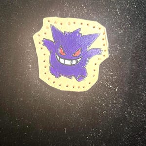 May include: A purple cartoon character with red eyes and a wide grin, set against a light tan background with small, evenly spaced holes around the edge. The character is a stylized illustration.