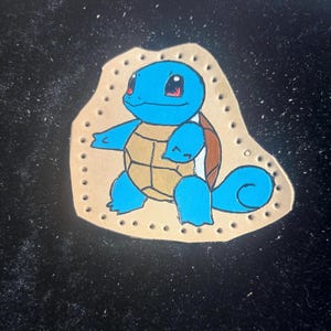 Puede incluir: Imagen de dibujos animados pintada a mano de un personaje Squirtle azul, con un caparazón marrón y detalles blancos. La imagen está sobre un fondo beige similar al cuero, con pequeños agujeros espaciados uniformemente alrededor del borde. El fondo está sobre una superficie negra.