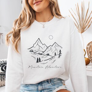 May include: White sweatshirt featuring a black mountain range graphic with a sun and the words "Mountain Adventure". The design includes trees and rolling hills. The sweatshirt is a crew neck style.
