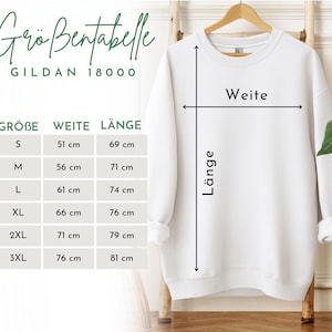 May include: A white Gildan 18000 sweatshirt displayed on a wooden hanger. The image includes a size chart with measurements in centimeters for width and length, ranging from size S to 3XL. The chart is labeled with German terms.