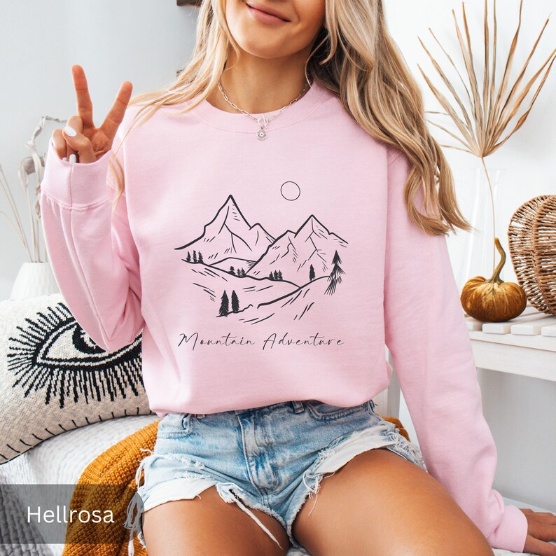 May include: A light pink sweatshirt with a black mountain graphic and the words "Mountain Adventure". The design features a mountain range, trees, and a sun. The sweatshirt is a crew neck style.