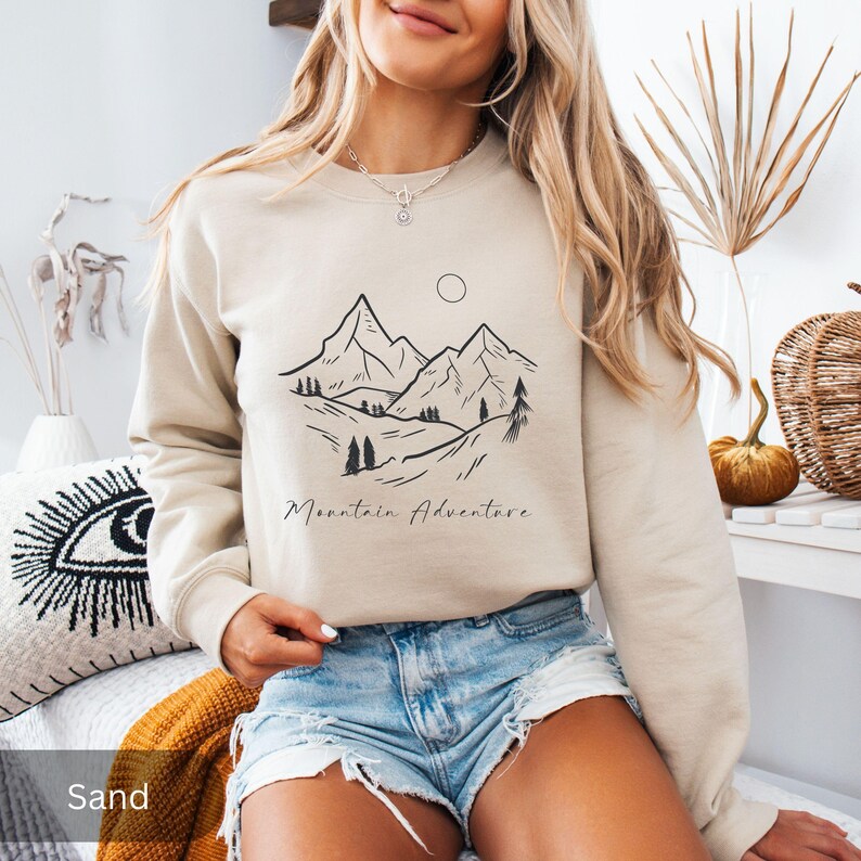 May include: Sand-colored sweatshirt with a black mountain graphic and the words "Mountain Adventure." The design features a mountain range, trees, and a sun. The sweatshirt is a neutral color.