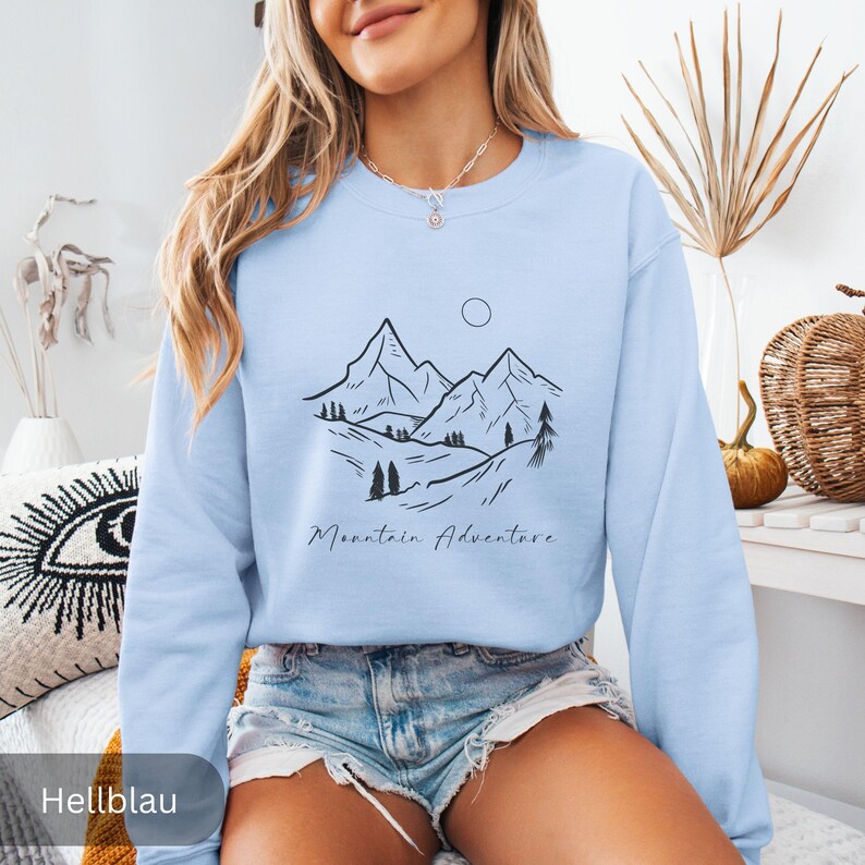 May include: Light blue sweatshirt with a black mountain landscape design and the words "Mountain Adventure" in a script font. The design includes mountains, trees, and a sun. The sweatshirt is a solid light blue color.