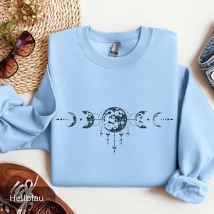 Könnte beinhalten: Ein hellblaues Sweatshirt mit einem schwarzen Motiv aus Mondphasen. Das Design zeigt einen Vollmond mit einer Mondsichel auf jeder Seite.