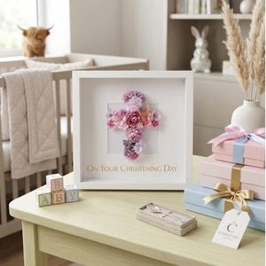 May include: A white framed floral cross with the text "On Your Christening Day". The cross is made of pink and purple flowers. Beside the frame are gift boxes, baby blocks, and a silver spoon set.