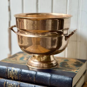 May include: A polished copper bowl with a wide rim, a rounded body, and a pedestal base. The bowl features two small, decorative metal handles attached to opposite sides. The surface of the copper has a reflective, metallic finish throughout.