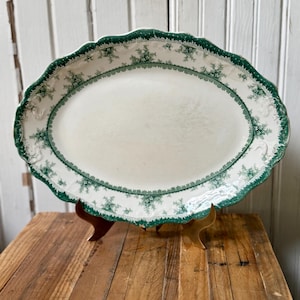May include: An oval ceramic serving platter featuring a cream-colored center and a decorative scalloped edge. The rim is adorned with a detailed green floral and geometric border pattern, providing a distinct contrast against the light base.