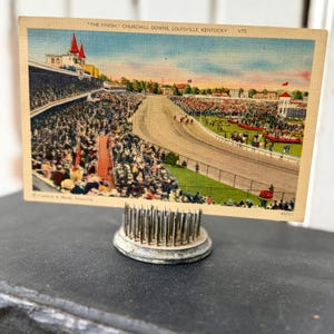 May include: Vintage postcard of Churchill Downs, Louisville, Kentucky, with the text "THE FINISH." The postcard shows a horse race with a packed grandstand and a red-roofed building. The postcard is held by a silver metal flower frog.