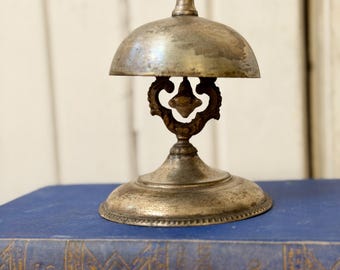 May include: A vintage silver-toned bell with a domed top and ornate base. The bell sits atop a blue book, showcasing its antique design. The bell's handle and decorative accents add to its classic appeal, perfect for a desk or counter.