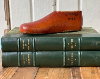 May include: A vintage, reddish-brown wooden shoe last, marked "STUART 5-55", rests atop two green hardcover books. The books have gold lettering and the titles "FEDERICO DE ROBERTO" and "LES VICE-ROIS". The background is a weathered, white-painted wall.