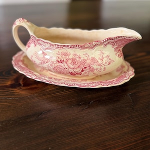 May include: A cream-colored ceramic gravy boat with an attached oval underplate, featuring a detailed red transferware pattern of floral motifs and birds. The piece has a scalloped rim, a curved handle, and a pouring spout with a glossy finish.