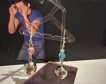 May include: A pair of dangle earrings featuring silver-toned disco ball charms, small silver star beads, and translucent faceted beads in pink and blue. The earrings are suspended from silver-toned metal hooks on a clear acrylic hanger display.
