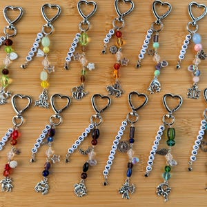May include: A collection of beaded zodiac keychains with silver-tone heart-shaped clasps. Each keychain features a different zodiac sign spelled out in white letter beads, along with colorful beads and charms. The keychains are displayed on a wooden surface.