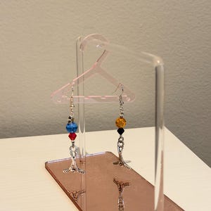 May include: Earrings with colorful beads, displayed on a pink acrylic hanger-shaped stand. One earring features blue and red beads, the other has yellow and black beads. The stand has a mirrored base.