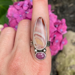 Puede incluir: Un anillo de plata con una piedra grande de forma ovalada con bandas crema, marrón y blancas. Debajo de la piedra principal hay una piedra preciosa rosa ovalada y facetada más pequeña. El anillo tiene detalles florales y de remolino decorativos.