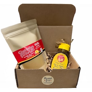 Puede incluir: Una caja de cartón que contiene una bolsa beige etiquetada como "Maria's Best Golden Milk" y una botella con una etiqueta amarilla. La botella tiene una tapa amarilla y contiene un líquido. La caja también incluye papel decorativo y una pegatina de "Thank You".
