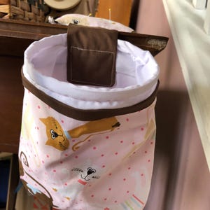 May include: A pink fabric storage bin with a brown fabric flap and a white lining. The bin has a repeating pattern of cartoon cats with pink polka dots.