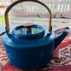 May include: A deep blue teapot with a smooth, glossy finish, featuring a circular lid with a small wooden knob handle. The teapot has a curved wooden handle attached to the sides with metal hardware and a short, angled spout on the right side.