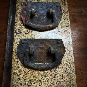 May include: Close-up of a vintage book with two antique metal handles. The book cover features a marbled pattern in yellow, brown, and red. The handles are dark, with a rustic, aged appearance, and are attached with rivets.