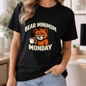 Puede incluir: Camiseta negra con el texto "BEAR MINIMUM MONDAY" y un oso de dibujos animados sosteniendo una taza de café. El oso es naranja y marrón con una expresión cansada. La camiseta la lleva una persona con vaqueros azules.